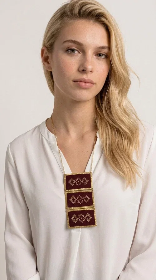 , The statement embroidered necklace in burgundy with geometric patterns is a piece of jewelry that combines the traditional art of embroidery with a modern aesthetic, creating a result that immediately catches the eye. Designed by Greek Jewelry Designer Aikaterini Chalkiadaki, this striking necklace draws inspiration from folk tradition and the geometric motifs that characterize Greek handicraft. The deep burgundy color of the fabric, combined with gold brass, creates a rich, warm palette that suits every skin tone.

The necklace consists of three rectangular embroidered sections, placed one below the other and framed with gold brass featuring a textured finish. Each section showcases handmade embroidery with repeating geometric patterns in a folklore style, reminiscent of traditional Greek embroidery. The result is a long necklace with distinctive character, ideal for elevating a simple outfit or completing a boho-chic look.