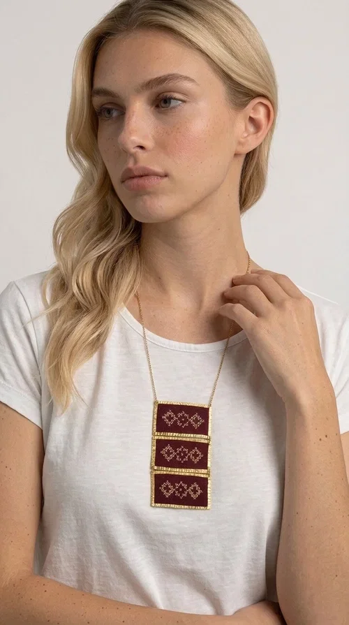 , The statement embroidered necklace in burgundy with geometric patterns is a piece of jewelry that combines the traditional art of embroidery with a modern aesthetic, creating a result that immediately catches the eye. Designed by Greek Jewelry Designer Aikaterini Chalkiadaki, this striking necklace draws inspiration from folk tradition and the geometric motifs that characterize Greek handicraft. The deep burgundy color of the fabric, combined with gold brass, creates a rich, warm palette that suits every skin tone.

The necklace consists of three rectangular embroidered sections, placed one below the other and framed with gold brass featuring a textured finish. Each section showcases handmade embroidery with repeating geometric patterns in a folklore style, reminiscent of traditional Greek embroidery. The result is a long necklace with distinctive character, ideal for elevating a simple outfit or completing a boho-chic look.