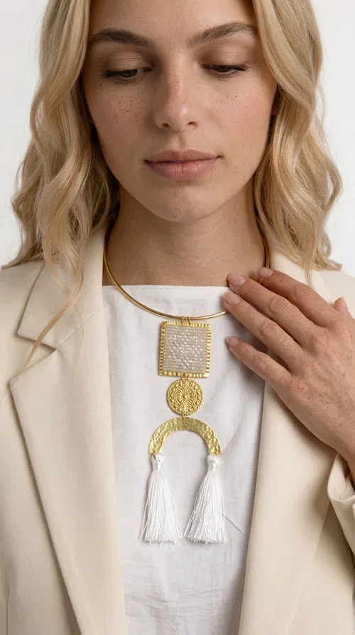 , A statement embroidered necklace that combines folk aesthetics with modern elegance — the ideal piece for those who love style with character.

Designed by Greek Jewelry Designer Aikaterini Chalkiadaki, this handmade necklace consists of three main elements: a rectangular embroidered pendant with geometric patterns in cream tones, a gold circular disc with embossed decoration, and a semicircular gold base adorned with white fabric fringes. The necklace is worn on a gold stainless steel omega chain that fits comfortably around the neck, adding balance to the striking pendant.

The materials used — brass and fabric — create a unique result that resembles a work of art. The geometric embroidered patterns are reminiscent of traditional Greek techniques, while the overall composition has a distinctly contemporary character. Ideal for casual chic looks as well as evening outings, it pairs perfectly with a white t-shirt, an oversized jacket, or a simple dress.