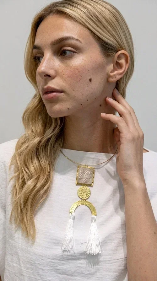 , A statement embroidered necklace that combines folk aesthetics with modern elegance — the ideal piece for those who love style with character.

Designed by Greek Jewelry Designer Aikaterini Chalkiadaki, this handmade necklace consists of three main elements: a rectangular embroidered pendant with geometric patterns in cream tones, a gold circular disc with embossed decoration, and a semicircular gold base adorned with white fabric fringes. The necklace is worn on a gold stainless steel omega chain that fits comfortably around the neck, adding balance to the striking pendant.

The materials used — brass and fabric — create a unique result that resembles a work of art. The geometric embroidered patterns are reminiscent of traditional Greek techniques, while the overall composition has a distinctly contemporary character. Ideal for casual chic looks as well as evening outings, it pairs perfectly with a white t-shirt, an oversized jacket, or a simple dress.