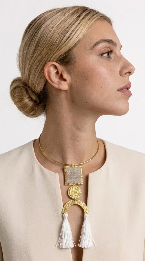 , A statement embroidered necklace that combines folk aesthetics with modern elegance — the ideal piece for those who love style with character.

Designed by Greek Jewelry Designer Aikaterini Chalkiadaki, this handmade necklace consists of three main elements: a rectangular embroidered pendant with geometric patterns in cream tones, a gold circular disc with embossed decoration, and a semicircular gold base adorned with white fabric fringes. The necklace is worn on a gold stainless steel omega chain that fits comfortably around the neck, adding balance to the striking pendant.

The materials used — brass and fabric — create a unique result that resembles a work of art. The geometric embroidered patterns are reminiscent of traditional Greek techniques, while the overall composition has a distinctly contemporary character. Ideal for casual chic looks as well as evening outings, it pairs perfectly with a white t-shirt, an oversized jacket, or a simple dress.