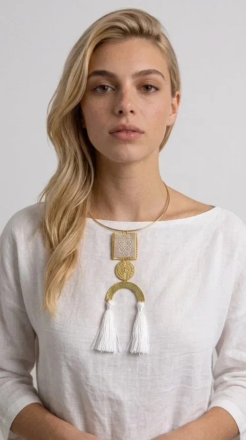 , A statement embroidered necklace that combines folk aesthetics with modern elegance — the ideal piece for those who love style with character.

Designed by Greek Jewelry Designer Aikaterini Chalkiadaki, this handmade necklace consists of three main elements: a rectangular embroidered pendant with geometric patterns in cream tones, a gold circular disc with embossed decoration, and a semicircular gold base adorned with white fabric fringes. The necklace is worn on a gold stainless steel omega chain that fits comfortably around the neck, adding balance to the striking pendant.

The materials used — brass and fabric — create a unique result that resembles a work of art. The geometric embroidered patterns are reminiscent of traditional Greek techniques, while the overall composition has a distinctly contemporary character. Ideal for casual chic looks as well as evening outings, it pairs perfectly with a white t-shirt, an oversized jacket, or a simple dress.