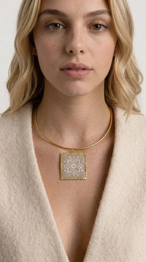 , An embroidered statement necklace that blends craftsmanship with contemporary aesthetics—ideal for those seeking jewelry with character and uniqueness.

Designed by Greek jewelry designer Aikaterini Chalkiadaki, this striking necklace features a square embroidered pendant in a cream hue, adorned with delicate geometric patterns that evoke the traditions of Greek folk art. The pendant rests on a gold-toned brass collar, giving the overall piece a structured, modern silhouette that pairs equally well with casual and formal attire.

The fabric's weave is dense and meticulous, featuring textured embroidered motifs that create a folklore-inspired aesthetic. The gold-toned brass frame encases the pendant with geometric precision, highlighting the handcrafted finesse of every detail.