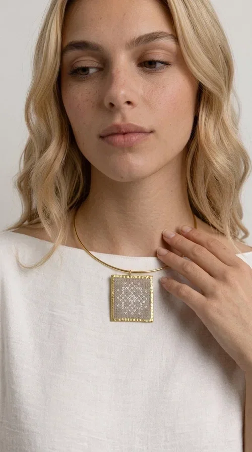 , An embroidered statement necklace that blends craftsmanship with contemporary aesthetics—ideal for those seeking jewelry with character and uniqueness.

Designed by Greek jewelry designer Aikaterini Chalkiadaki, this striking necklace features a square embroidered pendant in a cream hue, adorned with delicate geometric patterns that evoke the traditions of Greek folk art. The pendant rests on a gold-toned brass collar, giving the overall piece a structured, modern silhouette that pairs equally well with casual and formal attire.

The fabric's weave is dense and meticulous, featuring textured embroidered motifs that create a folklore-inspired aesthetic. The gold-toned brass frame encases the pendant with geometric precision, highlighting the handcrafted finesse of every detail.