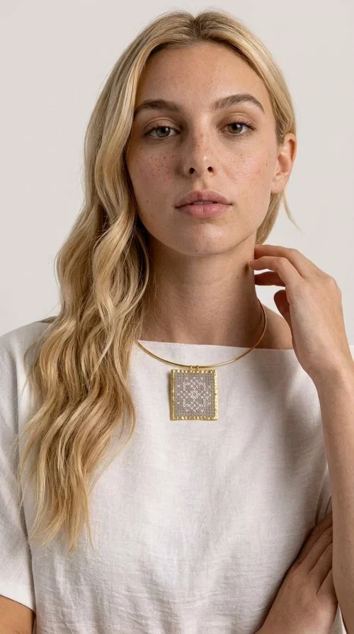 , An embroidered statement necklace that blends craftsmanship with contemporary aesthetics—ideal for those seeking jewelry with character and uniqueness.

Designed by Greek jewelry designer Aikaterini Chalkiadaki, this striking necklace features a square embroidered pendant in a cream hue, adorned with delicate geometric patterns that evoke the traditions of Greek folk art. The pendant rests on a gold-toned brass collar, giving the overall piece a structured, modern silhouette that pairs equally well with casual and formal attire.

The fabric's weave is dense and meticulous, featuring textured embroidered motifs that create a folklore-inspired aesthetic. The gold-toned brass frame encases the pendant with geometric precision, highlighting the handcrafted finesse of every detail.