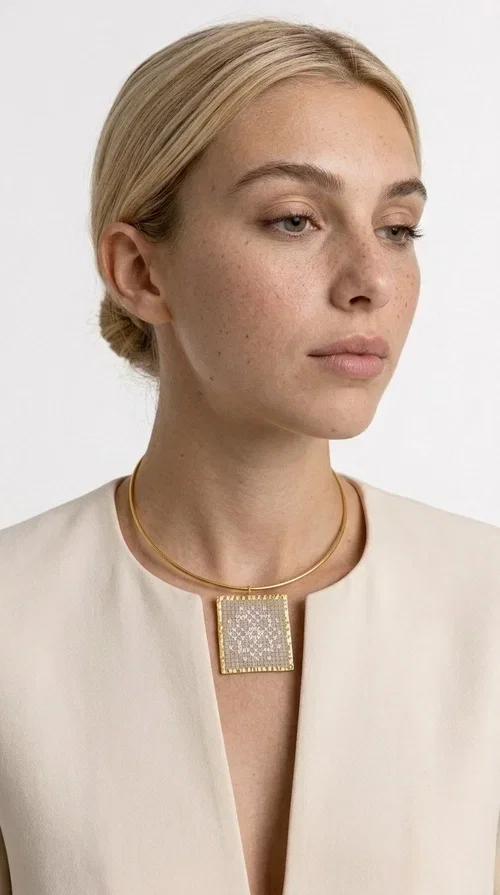 , An embroidered statement necklace that blends craftsmanship with contemporary aesthetics—ideal for those seeking jewelry with character and uniqueness.

Designed by Greek jewelry designer Aikaterini Chalkiadaki, this striking necklace features a square embroidered pendant in a cream hue, adorned with delicate geometric patterns that evoke the traditions of Greek folk art. The pendant rests on a gold-toned brass collar, giving the overall piece a structured, modern silhouette that pairs equally well with casual and formal attire.

The fabric's weave is dense and meticulous, featuring textured embroidered motifs that create a folklore-inspired aesthetic. The gold-toned brass frame encases the pendant with geometric precision, highlighting the handcrafted finesse of every detail.