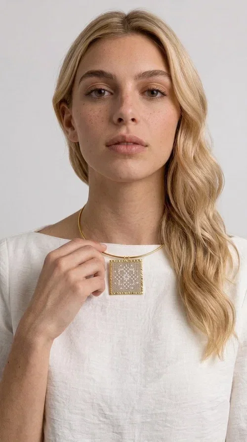 , An embroidered statement necklace that blends craftsmanship with contemporary aesthetics—ideal for those seeking jewelry with character and uniqueness.

Designed by Greek jewelry designer Aikaterini Chalkiadaki, this striking necklace features a square embroidered pendant in a cream hue, adorned with delicate geometric patterns that evoke the traditions of Greek folk art. The pendant rests on a gold-toned brass collar, giving the overall piece a structured, modern silhouette that pairs equally well with casual and formal attire.

The fabric's weave is dense and meticulous, featuring textured embroidered motifs that create a folklore-inspired aesthetic. The gold-toned brass frame encases the pendant with geometric precision, highlighting the handcrafted finesse of every detail.