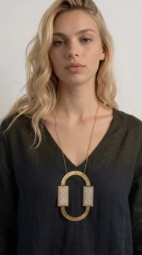 , A statement embroidered necklace that instantly catches the eye — featuring geometric patterns in an ecru shade combined with gold-toned brass that highlights its uniqueness. Designed by Greek Jewelry Designer Aikaterini Chalkiadaki, this handmade necklace blends the tradition of embroidery art with a modern, striking aesthetic result.

The ecru-colored embroidered fabric is set within two rectangular gold-toned brass frames, connected through a large oval element — creating a geometric composition full of character. The necklace hangs from a delicate gold chain, positioning the pendant right at the center of the chest for maximum visual impact. Ideal as a long statement necklace for casual or formal looks.

It pairs perfectly with simple dresses, pant-and-top sets, or low-cut tops that let the jewelry speak for itself. A perfect gift for loved ones who appreciate handmade, one-of-a-kind jewelry.