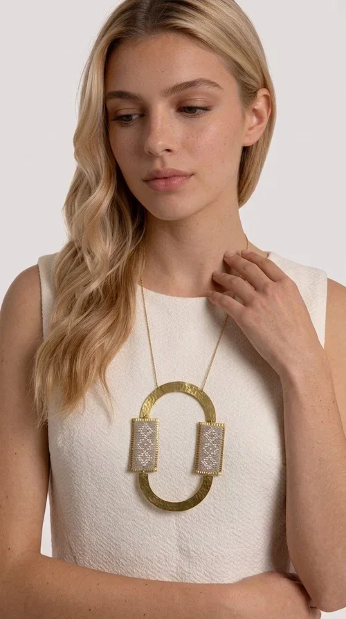 , A statement embroidered necklace that instantly catches the eye — featuring geometric patterns in an ecru shade combined with gold-toned brass that highlights its uniqueness. Designed by Greek Jewelry Designer Aikaterini Chalkiadaki, this handmade necklace blends the tradition of embroidery art with a modern, striking aesthetic result.

The ecru-colored embroidered fabric is set within two rectangular gold-toned brass frames, connected through a large oval element — creating a geometric composition full of character. The necklace hangs from a delicate gold chain, positioning the pendant right at the center of the chest for maximum visual impact. Ideal as a long statement necklace for casual or formal looks.

It pairs perfectly with simple dresses, pant-and-top sets, or low-cut tops that let the jewelry speak for itself. A perfect gift for loved ones who appreciate handmade, one-of-a-kind jewelry.
