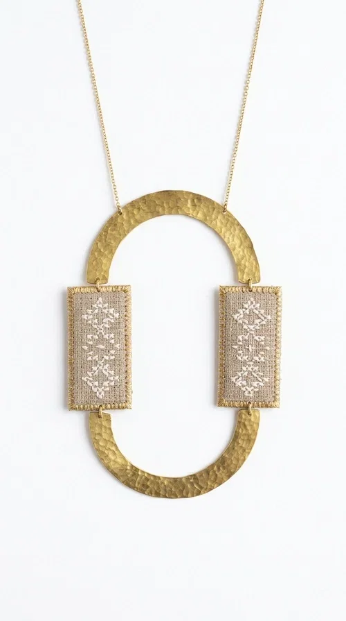 , A statement embroidered necklace that instantly catches the eye — featuring geometric patterns in an ecru shade combined with gold-toned brass that highlights its uniqueness. Designed by Greek Jewelry Designer Aikaterini Chalkiadaki, this handmade necklace blends the tradition of embroidery art with a modern, striking aesthetic result.

The ecru-colored embroidered fabric is set within two rectangular gold-toned brass frames, connected through a large oval element — creating a geometric composition full of character. The necklace hangs from a delicate gold chain, positioning the pendant right at the center of the chest for maximum visual impact. Ideal as a long statement necklace for casual or formal looks.

It pairs perfectly with simple dresses, pant-and-top sets, or low-cut tops that let the jewelry speak for itself. A perfect gift for loved ones who appreciate handmade, one-of-a-kind jewelry.