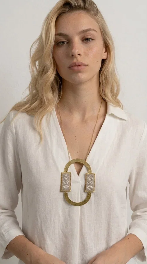 , A statement embroidered necklace that instantly catches the eye — featuring geometric patterns in an ecru shade combined with gold-toned brass that highlights its uniqueness. Designed by Greek Jewelry Designer Aikaterini Chalkiadaki, this handmade necklace blends the tradition of embroidery art with a modern, striking aesthetic result.

The ecru-colored embroidered fabric is set within two rectangular gold-toned brass frames, connected through a large oval element — creating a geometric composition full of character. The necklace hangs from a delicate gold chain, positioning the pendant right at the center of the chest for maximum visual impact. Ideal as a long statement necklace for casual or formal looks.

It pairs perfectly with simple dresses, pant-and-top sets, or low-cut tops that let the jewelry speak for itself. A perfect gift for loved ones who appreciate handmade, one-of-a-kind jewelry.