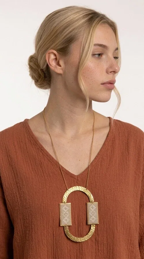 , A statement embroidered necklace that instantly catches the eye — featuring geometric patterns in an ecru shade combined with gold-toned brass that highlights its uniqueness. Designed by Greek Jewelry Designer Aikaterini Chalkiadaki, this handmade necklace blends the tradition of embroidery art with a modern, striking aesthetic result.

The ecru-colored embroidered fabric is set within two rectangular gold-toned brass frames, connected through a large oval element — creating a geometric composition full of character. The necklace hangs from a delicate gold chain, positioning the pendant right at the center of the chest for maximum visual impact. Ideal as a long statement necklace for casual or formal looks.

It pairs perfectly with simple dresses, pant-and-top sets, or low-cut tops that let the jewelry speak for itself. A perfect gift for loved ones who appreciate handmade, one-of-a-kind jewelry.