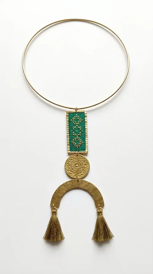, A statement embroidered necklace that instantly catches the eye — featuring a bold green color, geometric patterns and gold details reminiscent of the tradition of folk jewelry.

Designed by Greek Jewelry Designer Aikaterini Chalkiadaki, this handmade necklace combines brass and fabric in a composition that highlights the skill of Greek craftsmanship. The rectangular embroidered piece, with its delicate geometric motifs in gold and green tones, blends harmoniously with a gold circular disc and a brass arc, while three hanging tassels in a khaki-gold shade add movement and warmth to the overall look. It is worn on a gold rigid choker for an even more striking effect.

Ideal for looks that want to stand out — from summer evening outings to casual chic styles with simple linen or cotton clothing. It pairs perfectly with neutral tones, white, ecru, or deep green shades.