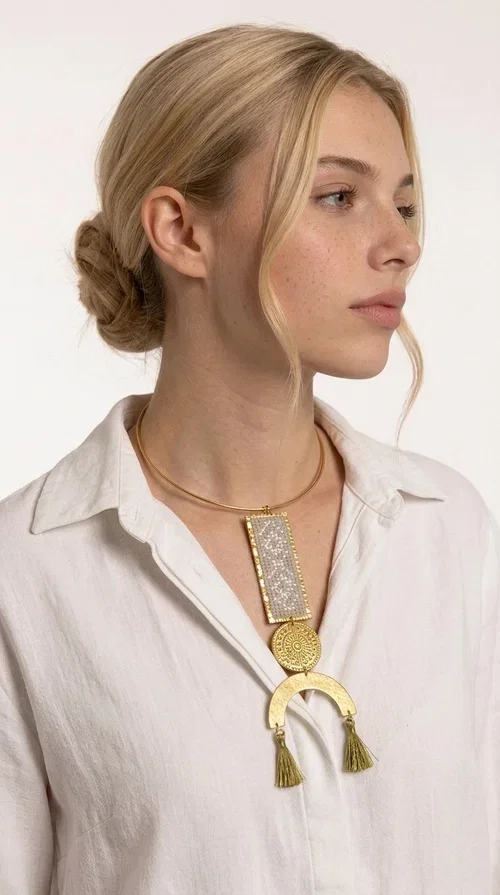 , A statement embroidered necklace that instantly catches the eye — featuring ivory geometric patterns on a brass frame, with details inspired by folklore tradition. Designed by Greek Jewelry Designer Aikaterini Chalkiadaki, this handmade necklace combines the art of embroidery with a modern aesthetic, creating a truly distinctive piece.

The necklace features a rectangular embroidered panel with delicate geometric motifs in shades of ivory and gold, a circular brass element with an embossed design at the center, and a semicircular base adorned with two gold tassels that add movement and a luxurious feel. The brass neck ring ensures a secure fit and timeless style.

Ideal for women who love striking jewelry with character, it pairs beautifully with simple ivory or white outfits, as well as bold colors for contrast. Perfect for evening outings, summer events, weddings, or stylish daytime looks.