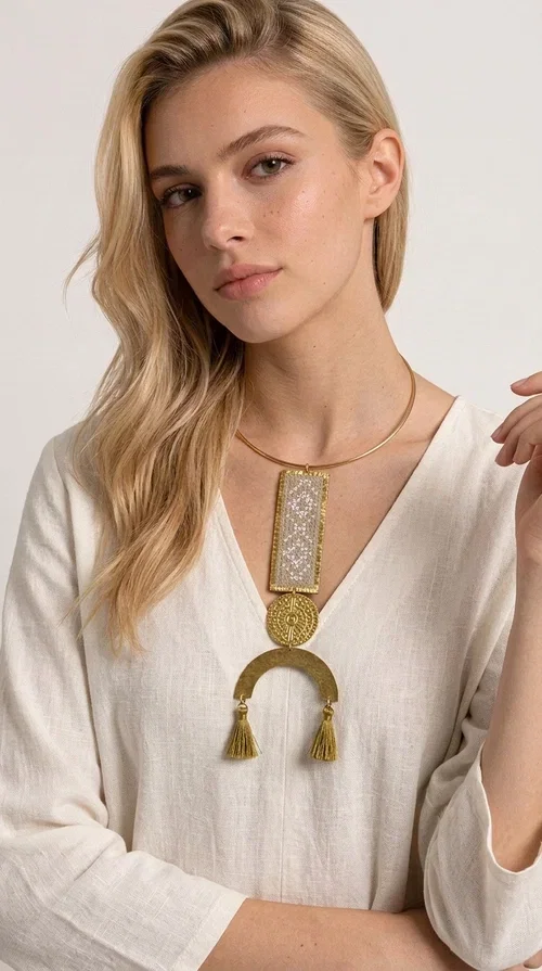, A statement embroidered necklace that instantly catches the eye — featuring ivory geometric patterns on a brass frame, with details inspired by folklore tradition. Designed by Greek Jewelry Designer Aikaterini Chalkiadaki, this handmade necklace combines the art of embroidery with a modern aesthetic, creating a truly distinctive piece.

The necklace features a rectangular embroidered panel with delicate geometric motifs in shades of ivory and gold, a circular brass element with an embossed design at the center, and a semicircular base adorned with two gold tassels that add movement and a luxurious feel. The brass neck ring ensures a secure fit and timeless style.

Ideal for women who love striking jewelry with character, it pairs beautifully with simple ivory or white outfits, as well as bold colors for contrast. Perfect for evening outings, summer events, weddings, or stylish daytime looks.