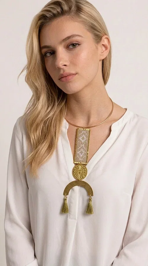 , A statement embroidered necklace that instantly catches the eye — featuring ivory geometric patterns on a brass frame, with details inspired by folklore tradition. Designed by Greek Jewelry Designer Aikaterini Chalkiadaki, this handmade necklace combines the art of embroidery with a modern aesthetic, creating a truly distinctive piece.

The necklace features a rectangular embroidered panel with delicate geometric motifs in shades of ivory and gold, a circular brass element with an embossed design at the center, and a semicircular base adorned with two gold tassels that add movement and a luxurious feel. The brass neck ring ensures a secure fit and timeless style.

Ideal for women who love striking jewelry with character, it pairs beautifully with simple ivory or white outfits, as well as bold colors for contrast. Perfect for evening outings, summer events, weddings, or stylish daytime looks.