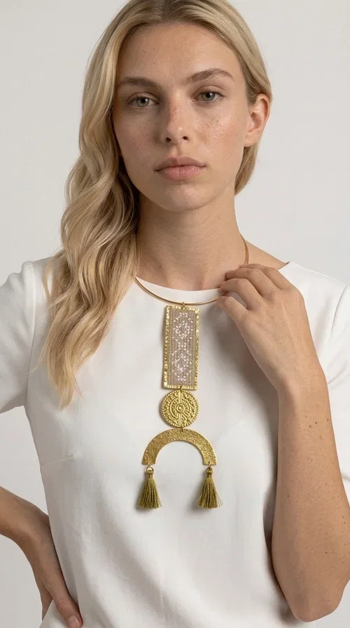 , A statement embroidered necklace that instantly catches the eye — featuring ivory geometric patterns on a brass frame, with details inspired by folklore tradition. Designed by Greek Jewelry Designer Aikaterini Chalkiadaki, this handmade necklace combines the art of embroidery with a modern aesthetic, creating a truly distinctive piece.

The necklace features a rectangular embroidered panel with delicate geometric motifs in shades of ivory and gold, a circular brass element with an embossed design at the center, and a semicircular base adorned with two gold tassels that add movement and a luxurious feel. The brass neck ring ensures a secure fit and timeless style.

Ideal for women who love striking jewelry with character, it pairs beautifully with simple ivory or white outfits, as well as bold colors for contrast. Perfect for evening outings, summer events, weddings, or stylish daytime looks.