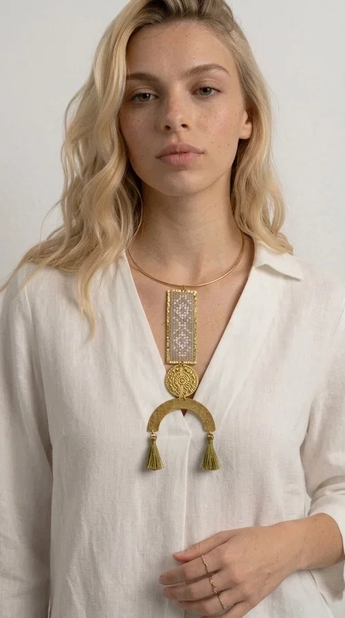 , A statement embroidered necklace that instantly catches the eye — featuring ivory geometric patterns on a brass frame, with details inspired by folklore tradition. Designed by Greek Jewelry Designer Aikaterini Chalkiadaki, this handmade necklace combines the art of embroidery with a modern aesthetic, creating a truly distinctive piece.

The necklace features a rectangular embroidered panel with delicate geometric motifs in shades of ivory and gold, a circular brass element with an embossed design at the center, and a semicircular base adorned with two gold tassels that add movement and a luxurious feel. The brass neck ring ensures a secure fit and timeless style.

Ideal for women who love striking jewelry with character, it pairs beautifully with simple ivory or white outfits, as well as bold colors for contrast. Perfect for evening outings, summer events, weddings, or stylish daytime looks.