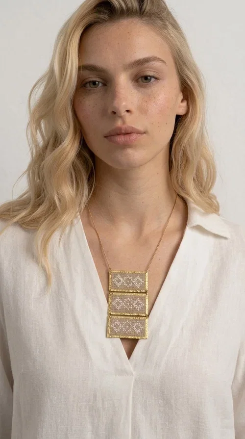 , The statement embroidered ivory necklace is a handmade creation that combines the traditional art of embroidery with a contemporary jewelry aesthetic, adding a unique, eye-catching touch to any outfit.

Designed by Greek Jewelry Designer Aikaterini Chalkiadaki, this necklace features three rectangular embroidered sections with ivory geometric motifs and gold details set in a brass frame. It hangs from a delicate gold chain that lends elegance and lightness, while the folklore-style geometric patterns give the piece a distinctive, ethnographic character. The combination of brass and fabric creates an intriguing contrast of materials that draws the eye.

Ideal for both day and evening wear, it pairs beautifully with white or monochrome outfits to highlight its geometric patterns. The ivory color makes it versatile and timeless, perfect for summer looks as well as formal or business occasions.