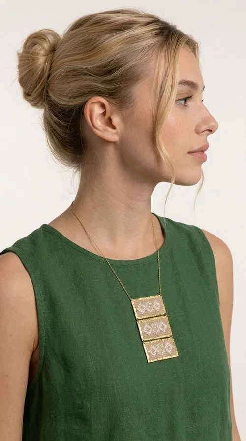 , The statement embroidered ivory necklace is a handmade creation that combines the traditional art of embroidery with a contemporary jewelry aesthetic, adding a unique, eye-catching touch to any outfit.

Designed by Greek Jewelry Designer Aikaterini Chalkiadaki, this necklace features three rectangular embroidered sections with ivory geometric motifs and gold details set in a brass frame. It hangs from a delicate gold chain that lends elegance and lightness, while the folklore-style geometric patterns give the piece a distinctive, ethnographic character. The combination of brass and fabric creates an intriguing contrast of materials that draws the eye.

Ideal for both day and evening wear, it pairs beautifully with white or monochrome outfits to highlight its geometric patterns. The ivory color makes it versatile and timeless, perfect for summer looks as well as formal or business occasions.