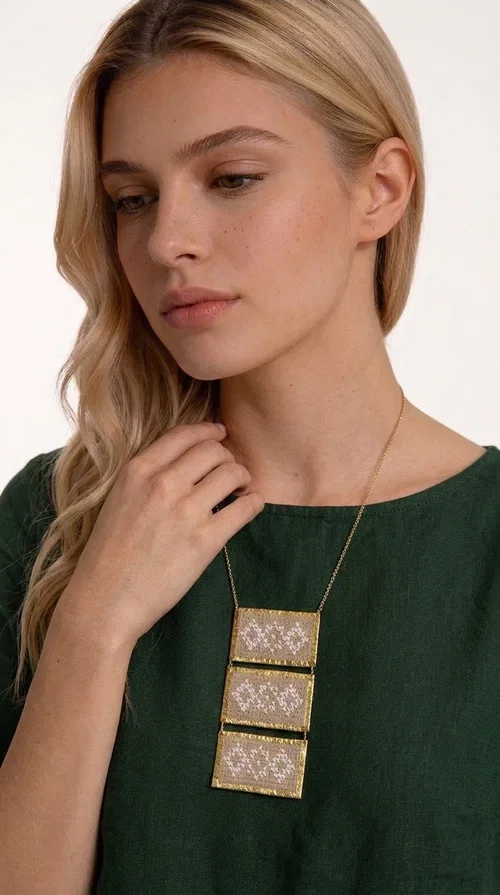 , The statement embroidered ivory necklace is a handmade creation that combines the traditional art of embroidery with a contemporary jewelry aesthetic, adding a unique, eye-catching touch to any outfit.

Designed by Greek Jewelry Designer Aikaterini Chalkiadaki, this necklace features three rectangular embroidered sections with ivory geometric motifs and gold details set in a brass frame. It hangs from a delicate gold chain that lends elegance and lightness, while the folklore-style geometric patterns give the piece a distinctive, ethnographic character. The combination of brass and fabric creates an intriguing contrast of materials that draws the eye.

Ideal for both day and evening wear, it pairs beautifully with white or monochrome outfits to highlight its geometric patterns. The ivory color makes it versatile and timeless, perfect for summer looks as well as formal or business occasions.