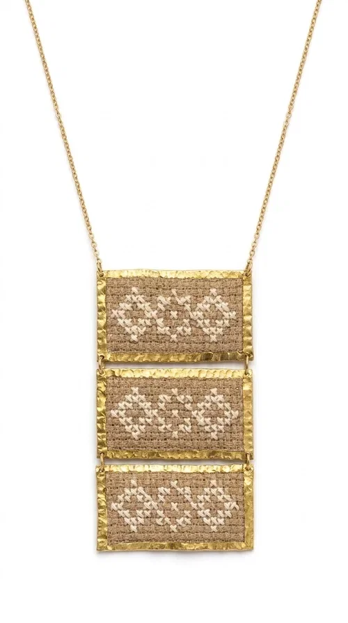 , The statement embroidered ivory necklace is a handmade creation that combines the traditional art of embroidery with a contemporary jewelry aesthetic, adding a unique, eye-catching touch to any outfit.

Designed by Greek Jewelry Designer Aikaterini Chalkiadaki, this necklace features three rectangular embroidered sections with ivory geometric motifs and gold details set in a brass frame. It hangs from a delicate gold chain that lends elegance and lightness, while the folklore-style geometric patterns give the piece a distinctive, ethnographic character. The combination of brass and fabric creates an intriguing contrast of materials that draws the eye.

Ideal for both day and evening wear, it pairs beautifully with white or monochrome outfits to highlight its geometric patterns. The ivory color makes it versatile and timeless, perfect for summer looks as well as formal or business occasions.
