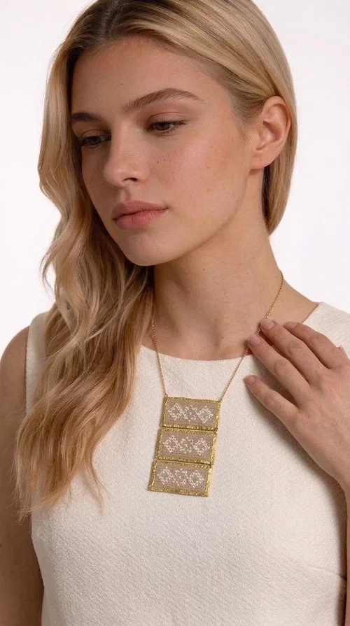 , The statement embroidered ivory necklace is a handmade creation that combines the traditional art of embroidery with a contemporary jewelry aesthetic, adding a unique, eye-catching touch to any outfit.

Designed by Greek Jewelry Designer Aikaterini Chalkiadaki, this necklace features three rectangular embroidered sections with ivory geometric motifs and gold details set in a brass frame. It hangs from a delicate gold chain that lends elegance and lightness, while the folklore-style geometric patterns give the piece a distinctive, ethnographic character. The combination of brass and fabric creates an intriguing contrast of materials that draws the eye.

Ideal for both day and evening wear, it pairs beautifully with white or monochrome outfits to highlight its geometric patterns. The ivory color makes it versatile and timeless, perfect for summer looks as well as formal or business occasions.