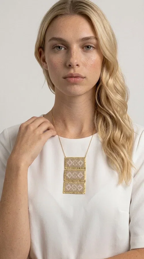 , The statement embroidered ivory necklace is a handmade creation that combines the traditional art of embroidery with a contemporary jewelry aesthetic, adding a unique, eye-catching touch to any outfit.

Designed by Greek Jewelry Designer Aikaterini Chalkiadaki, this necklace features three rectangular embroidered sections with ivory geometric motifs and gold details set in a brass frame. It hangs from a delicate gold chain that lends elegance and lightness, while the folklore-style geometric patterns give the piece a distinctive, ethnographic character. The combination of brass and fabric creates an intriguing contrast of materials that draws the eye.

Ideal for both day and evening wear, it pairs beautifully with white or monochrome outfits to highlight its geometric patterns. The ivory color makes it versatile and timeless, perfect for summer looks as well as formal or business occasions.