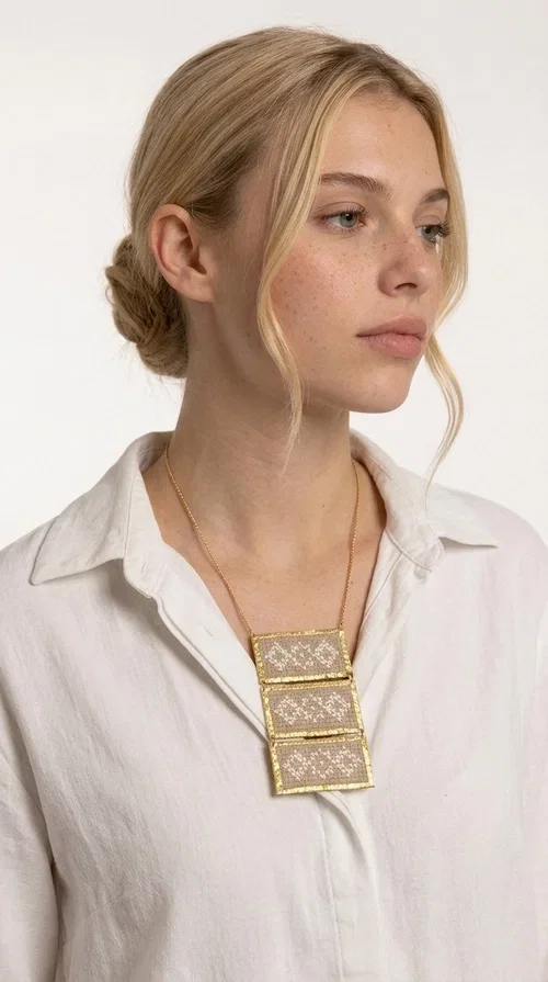 , The statement embroidered ivory necklace is a handmade creation that combines the traditional art of embroidery with a contemporary jewelry aesthetic, adding a unique, eye-catching touch to any outfit.

Designed by Greek Jewelry Designer Aikaterini Chalkiadaki, this necklace features three rectangular embroidered sections with ivory geometric motifs and gold details set in a brass frame. It hangs from a delicate gold chain that lends elegance and lightness, while the folklore-style geometric patterns give the piece a distinctive, ethnographic character. The combination of brass and fabric creates an intriguing contrast of materials that draws the eye.

Ideal for both day and evening wear, it pairs beautifully with white or monochrome outfits to highlight its geometric patterns. The ivory color makes it versatile and timeless, perfect for summer looks as well as formal or business occasions.