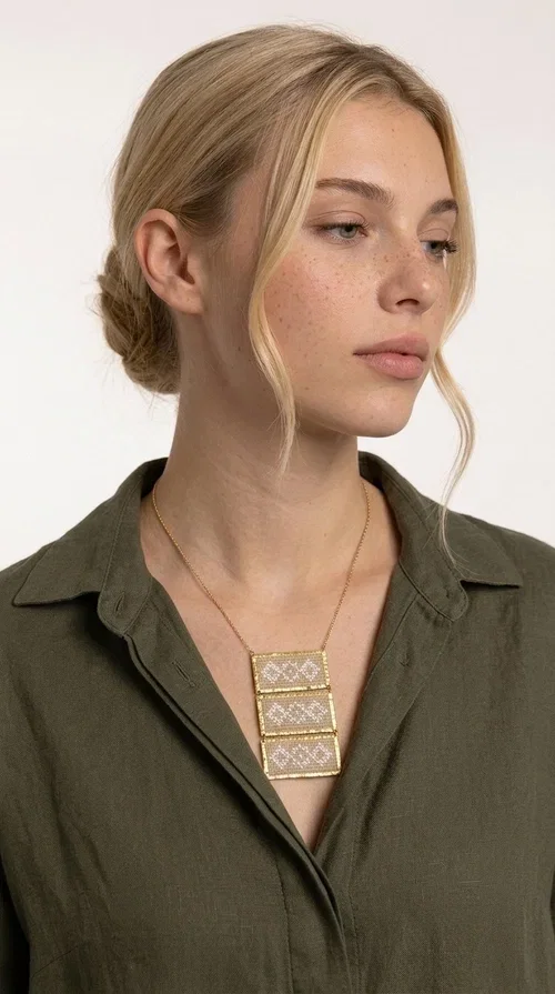 , The statement embroidered ivory necklace is a handmade creation that combines the traditional art of embroidery with a contemporary jewelry aesthetic, adding a unique, eye-catching touch to any outfit.

Designed by Greek Jewelry Designer Aikaterini Chalkiadaki, this necklace features three rectangular embroidered sections with ivory geometric motifs and gold details set in a brass frame. It hangs from a delicate gold chain that lends elegance and lightness, while the folklore-style geometric patterns give the piece a distinctive, ethnographic character. The combination of brass and fabric creates an intriguing contrast of materials that draws the eye.

Ideal for both day and evening wear, it pairs beautifully with white or monochrome outfits to highlight its geometric patterns. The ivory color makes it versatile and timeless, perfect for summer looks as well as formal or business occasions.
