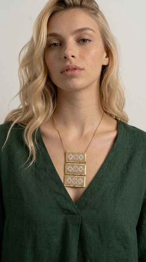 , The statement embroidered ivory necklace is a handmade creation that combines the traditional art of embroidery with a contemporary jewelry aesthetic, adding a unique, eye-catching touch to any outfit.

Designed by Greek Jewelry Designer Aikaterini Chalkiadaki, this necklace features three rectangular embroidered sections with ivory geometric motifs and gold details set in a brass frame. It hangs from a delicate gold chain that lends elegance and lightness, while the folklore-style geometric patterns give the piece a distinctive, ethnographic character. The combination of brass and fabric creates an intriguing contrast of materials that draws the eye.

Ideal for both day and evening wear, it pairs beautifully with white or monochrome outfits to highlight its geometric patterns. The ivory color makes it versatile and timeless, perfect for summer looks as well as formal or business occasions.