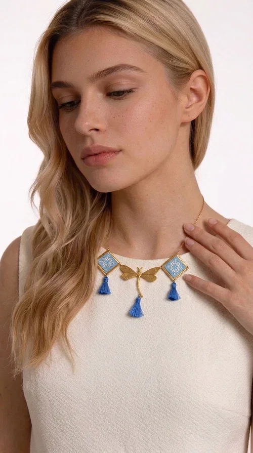 , A statement necklace that combines the tradition of embroidery art with contemporary aesthetics — ideal for those who want to stand out with style and character.

Designed by Greek Jewelry Designer Aikaterini Chalkiadaki, this handmade embroidered necklace in light blue brings to the forefront the magic of geometric patterns inspired by Greek folk tradition. Fabric rhombuses with finely worked embroidery in light blue and white shades alternate with a golden dragonfly made of brass, while four hanging tassels in deep blue add movement and vibrancy to the composition. The golden brass chain completes a creation full of color, texture and atmosphere.

Wear it over a white shirt or a simple monochrome dress to highlight all its beauty. It fits perfectly with summer looks, romantic evening outings, and boho-chic day looks as well. It is also an excellent gift choice for a woman who loves striking, handmade jewelry.