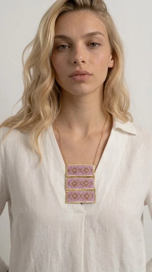 , An embroidered necklace that combines tradition with contemporary aesthetics — the Statement Embroidered Necklace in Lilac with Geometric Patterns is the jewel that turns any look into a style statement. Designed by Greek Jewelry Designer Aikaterini Chalkiadaki, this long necklace carries the distinctive energy of folklore aesthetics in a bold, striking version.

Three rectangular brass plates in a gold hue are covered with handmade embroidery in lilac and gold tones, featuring repeating geometric motifs inspired by Greek tradition. They hang from a delicate gold chain, creating a long necklace with a statement presence that suits both casual and elegant looks.

Ideal to pair with linen, cotton or evening wear, this necklace adds character to any outfit — from summer outings to formal occasions. The lilac hue is romantic and versatile, while the geometric patterns bring a strong personality.