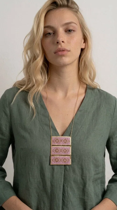 , An embroidered necklace that combines tradition with contemporary aesthetics — the Statement Embroidered Necklace in Lilac with Geometric Patterns is the jewel that turns any look into a style statement. Designed by Greek Jewelry Designer Aikaterini Chalkiadaki, this long necklace carries the distinctive energy of folklore aesthetics in a bold, striking version.

Three rectangular brass plates in a gold hue are covered with handmade embroidery in lilac and gold tones, featuring repeating geometric motifs inspired by Greek tradition. They hang from a delicate gold chain, creating a long necklace with a statement presence that suits both casual and elegant looks.

Ideal to pair with linen, cotton or evening wear, this necklace adds character to any outfit — from summer outings to formal occasions. The lilac hue is romantic and versatile, while the geometric patterns bring a strong personality.