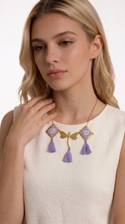, A statement embroidered necklace that blends the traditional art of embroidery with a contemporary aesthetic — ideal for every woman who wants to express her uniqueness with style.

Designed by Greek Jewelry Designer Aikaterini Chalkiadaki, this striking necklace features two diamond-shaped pendants with handmade embroidery in shades of lilac, a gold brass facing deer (libelle) charm at the center, and four fabric tassels in lilac tones that move gracefully with every step. The gold brass chain that fastens at the back of the neck gives the piece a delicate, elegant silhouette. The geometric embroidery patterns are reminiscent of authentic Greek folk motifs, evoking the rich tradition of Greek handicraft.

Perfect for summer looks, celebrations, or everyday moments that call for something special, this handmade necklace pairs beautifully with a white shirt, a dress, or a boho outfit. Pair it with simple earrings to let it stand out to the fullest.