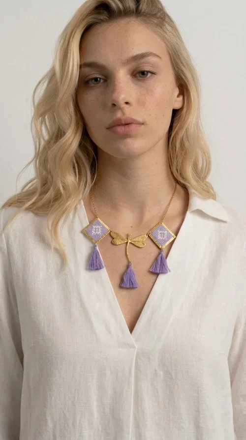 , A statement embroidered necklace that blends the traditional art of embroidery with a contemporary aesthetic — ideal for every woman who wants to express her uniqueness with style.

Designed by Greek Jewelry Designer Aikaterini Chalkiadaki, this striking necklace features two diamond-shaped pendants with handmade embroidery in shades of lilac, a gold brass facing deer (libelle) charm at the center, and four fabric tassels in lilac tones that move gracefully with every step. The gold brass chain that fastens at the back of the neck gives the piece a delicate, elegant silhouette. The geometric embroidery patterns are reminiscent of authentic Greek folk motifs, evoking the rich tradition of Greek handicraft.

Perfect for summer looks, celebrations, or everyday moments that call for something special, this handmade necklace pairs beautifully with a white shirt, a dress, or a boho outfit. Pair it with simple earrings to let it stand out to the fullest.