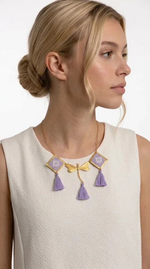 , A statement embroidered necklace that blends the traditional art of embroidery with a contemporary aesthetic — ideal for every woman who wants to express her uniqueness with style.

Designed by Greek Jewelry Designer Aikaterini Chalkiadaki, this striking necklace features two diamond-shaped pendants with handmade embroidery in shades of lilac, a gold brass facing deer (libelle) charm at the center, and four fabric tassels in lilac tones that move gracefully with every step. The gold brass chain that fastens at the back of the neck gives the piece a delicate, elegant silhouette. The geometric embroidery patterns are reminiscent of authentic Greek folk motifs, evoking the rich tradition of Greek handicraft.

Perfect for summer looks, celebrations, or everyday moments that call for something special, this handmade necklace pairs beautifully with a white shirt, a dress, or a boho outfit. Pair it with simple earrings to let it stand out to the fullest.