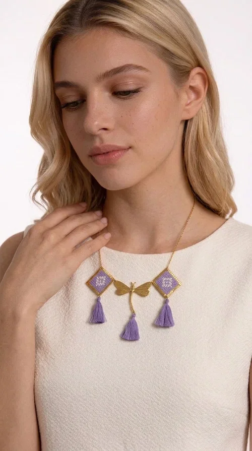 , A statement embroidered necklace that blends the traditional art of embroidery with a contemporary aesthetic — ideal for every woman who wants to express her uniqueness with style.

Designed by Greek Jewelry Designer Aikaterini Chalkiadaki, this striking necklace features two diamond-shaped pendants with handmade embroidery in shades of lilac, a gold brass facing deer (libelle) charm at the center, and four fabric tassels in lilac tones that move gracefully with every step. The gold brass chain that fastens at the back of the neck gives the piece a delicate, elegant silhouette. The geometric embroidery patterns are reminiscent of authentic Greek folk motifs, evoking the rich tradition of Greek handicraft.

Perfect for summer looks, celebrations, or everyday moments that call for something special, this handmade necklace pairs beautifully with a white shirt, a dress, or a boho outfit. Pair it with simple earrings to let it stand out to the fullest.