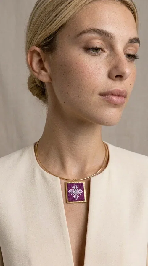 , The embroidered magenta necklace with geometric patterns is a statement piece that combines the traditional art of embroidery with a modern aesthetic — ideal for every woman who wants to express herself boldly and stylishly.

Designed by Greek Jewelry Designer Aikaterini Chalkiadaki, this handmade necklace features a square pendant in deep purple-magenta fabric, adorned with a white embroidered geometric design inspired by folk art tradition. The golden brass metal frame elegantly encases the pendant, while the delicate gold choker-style chain showcases it flawlessly around the neck. The result is a piece of jewelry with soul, character, and a strong visual presence.

Wear it over a white shirt, dress, or blazer to create a striking combination. It looks equally great in casual and more formal outfits, making it a versatile accessory for any occasion.