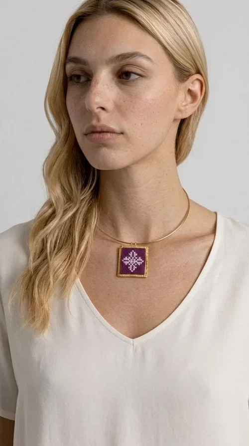 , The embroidered magenta necklace with geometric patterns is a statement piece that combines the traditional art of embroidery with a modern aesthetic — ideal for every woman who wants to express herself boldly and stylishly.

Designed by Greek Jewelry Designer Aikaterini Chalkiadaki, this handmade necklace features a square pendant in deep purple-magenta fabric, adorned with a white embroidered geometric design inspired by folk art tradition. The golden brass metal frame elegantly encases the pendant, while the delicate gold choker-style chain showcases it flawlessly around the neck. The result is a piece of jewelry with soul, character, and a strong visual presence.

Wear it over a white shirt, dress, or blazer to create a striking combination. It looks equally great in casual and more formal outfits, making it a versatile accessory for any occasion.