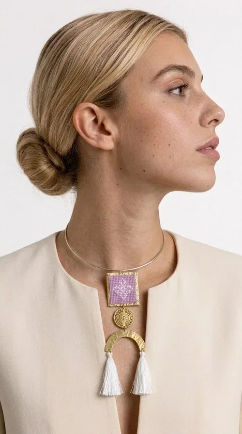 , A statement embroidered necklace that combines the traditional art of embroidery with a modern aesthetic — the ideal piece for every woman who wants to express herself with style and authenticity.

Designed by Greek Jewelry Designer Aikaterini Chalkiadaki, this handmade necklace features an embroidered square motif in pink/lilac tones with white geometric patterns inspired by Greek folk tradition. The embroidered piece is framed in golden brass and paired with a round embossed motif and a semicircular gold base, from which delicate white tassels hang, adding movement and grace. The look is completed with a fine gold choker-style chain.

Ideal to wear over a white top, a dress, or even a simple casual look that needs a touch of character. This striking long necklace turns any outfit into a true styling statement.