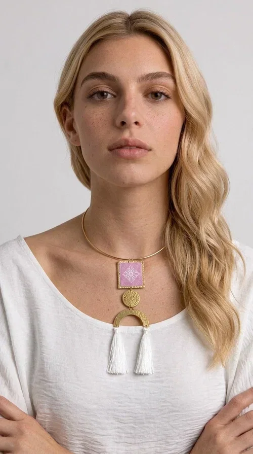 , A statement embroidered necklace that combines the traditional art of embroidery with a modern aesthetic — the ideal piece for every woman who wants to express herself with style and authenticity.

Designed by Greek Jewelry Designer Aikaterini Chalkiadaki, this handmade necklace features an embroidered square motif in pink/lilac tones with white geometric patterns inspired by Greek folk tradition. The embroidered piece is framed in golden brass and paired with a round embossed motif and a semicircular gold base, from which delicate white tassels hang, adding movement and grace. The look is completed with a fine gold choker-style chain.

Ideal to wear over a white top, a dress, or even a simple casual look that needs a touch of character. This striking long necklace turns any outfit into a true styling statement.
