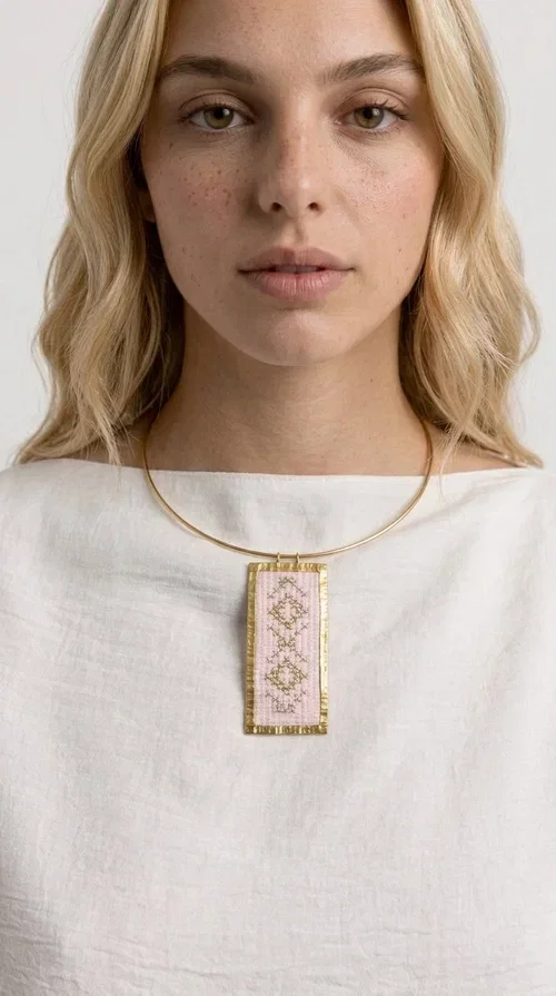 , The embroidered pink necklace with geometric patterns is a statement piece that combines the traditional art of embroidery with a modern, striking result. Designed by Greek Jewelry Designer Aikaterini Chalkiadaki, this unique necklace draws inspiration from folk tradition and the folk aesthetic that defines the Folklore Jewelry collection.

The embroidered rectangular pendant, in a soft pink shade, is adorned with gold geometric motifs reminiscent of traditional embroidery, while the gold brass metal frame gives it structure and elegance. It is worn on a delicate gold torque-style neck hoop that fits comfortably and instantly elevates any outfit, from casual to formal.

Ideal for women who love handmade jewelry with character, it is the perfect choice as a gift or to refresh your own jewelry collection with a piece full of soul.