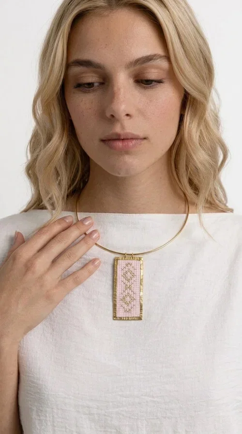 , The embroidered pink necklace with geometric patterns is a statement piece that combines the traditional art of embroidery with a modern, striking result. Designed by Greek Jewelry Designer Aikaterini Chalkiadaki, this unique necklace draws inspiration from folk tradition and the folk aesthetic that defines the Folklore Jewelry collection.

The embroidered rectangular pendant, in a soft pink shade, is adorned with gold geometric motifs reminiscent of traditional embroidery, while the gold brass metal frame gives it structure and elegance. It is worn on a delicate gold torque-style neck hoop that fits comfortably and instantly elevates any outfit, from casual to formal.

Ideal for women who love handmade jewelry with character, it is the perfect choice as a gift or to refresh your own jewelry collection with a piece full of soul.
