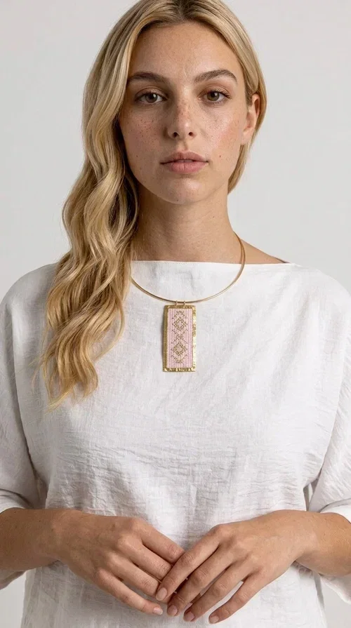 , The embroidered pink necklace with geometric patterns is a statement piece that combines the traditional art of embroidery with a modern, striking result. Designed by Greek Jewelry Designer Aikaterini Chalkiadaki, this unique necklace draws inspiration from folk tradition and the folk aesthetic that defines the Folklore Jewelry collection.

The embroidered rectangular pendant, in a soft pink shade, is adorned with gold geometric motifs reminiscent of traditional embroidery, while the gold brass metal frame gives it structure and elegance. It is worn on a delicate gold torque-style neck hoop that fits comfortably and instantly elevates any outfit, from casual to formal.

Ideal for women who love handmade jewelry with character, it is the perfect choice as a gift or to refresh your own jewelry collection with a piece full of soul.