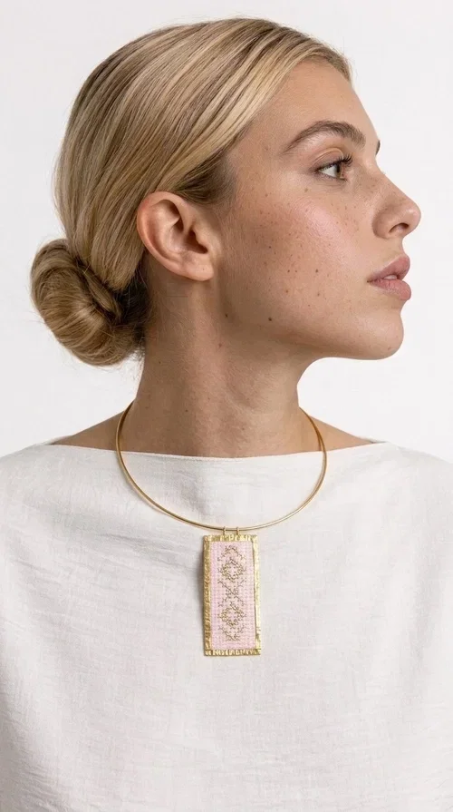 , The embroidered pink necklace with geometric patterns is a statement piece that combines the traditional art of embroidery with a modern, striking result. Designed by Greek Jewelry Designer Aikaterini Chalkiadaki, this unique necklace draws inspiration from folk tradition and the folk aesthetic that defines the Folklore Jewelry collection.

The embroidered rectangular pendant, in a soft pink shade, is adorned with gold geometric motifs reminiscent of traditional embroidery, while the gold brass metal frame gives it structure and elegance. It is worn on a delicate gold torque-style neck hoop that fits comfortably and instantly elevates any outfit, from casual to formal.

Ideal for women who love handmade jewelry with character, it is the perfect choice as a gift or to refresh your own jewelry collection with a piece full of soul.