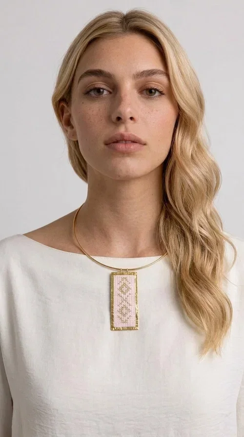 , The embroidered pink necklace with geometric patterns is a statement piece that combines the traditional art of embroidery with a modern, striking result. Designed by Greek Jewelry Designer Aikaterini Chalkiadaki, this unique necklace draws inspiration from folk tradition and the folk aesthetic that defines the Folklore Jewelry collection.

The embroidered rectangular pendant, in a soft pink shade, is adorned with gold geometric motifs reminiscent of traditional embroidery, while the gold brass metal frame gives it structure and elegance. It is worn on a delicate gold torque-style neck hoop that fits comfortably and instantly elevates any outfit, from casual to formal.

Ideal for women who love handmade jewelry with character, it is the perfect choice as a gift or to refresh your own jewelry collection with a piece full of soul.