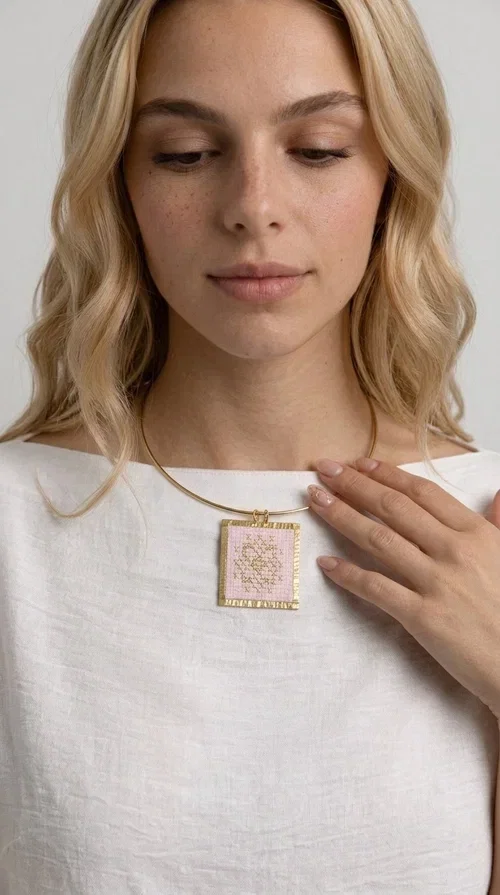, The statement embroidered necklace in a pink hue is a piece of jewelry that combines the traditional art of embroidery with contemporary design, creating a result that immediately catches the eye. Designed by Aikaterini Chalkiadaki, it carries the signature of Greek handmade craftsmanship and is an ideal choice for those seeking jewelry with character and history.

The necklace features a rectangular embroidered pendant with geometric patterns inspired by folk art and a folklore aesthetic. The pink fabric base combined with golden brass details creates a harmonious contrast that highlights the handmade element. The gold-toned metal omega-style chain gives structure and elegance to the whole, while the pendant is framed by distinctive golden details that emphasize the craftsmanship.

Wear it over a white t-shirt for a casual look or pair it with a blazer for a professional or evening look — just as shown in the photo. It is a piece of jewelry that suits every style and makes an excellent gift idea.
