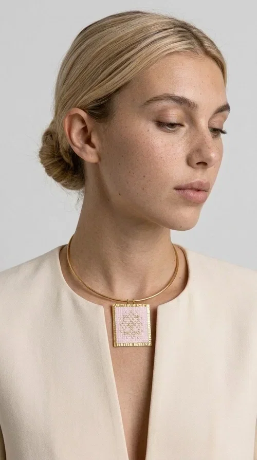 , The statement embroidered necklace in a pink hue is a piece of jewelry that combines the traditional art of embroidery with contemporary design, creating a result that immediately catches the eye. Designed by Aikaterini Chalkiadaki, it carries the signature of Greek handmade craftsmanship and is an ideal choice for those seeking jewelry with character and history.

The necklace features a rectangular embroidered pendant with geometric patterns inspired by folk art and a folklore aesthetic. The pink fabric base combined with golden brass details creates a harmonious contrast that highlights the handmade element. The gold-toned metal omega-style chain gives structure and elegance to the whole, while the pendant is framed by distinctive golden details that emphasize the craftsmanship.

Wear it over a white t-shirt for a casual look or pair it with a blazer for a professional or evening look — just as shown in the photo. It is a piece of jewelry that suits every style and makes an excellent gift idea.
