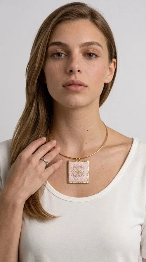 , The statement embroidered necklace in a pink hue is a piece of jewelry that combines the traditional art of embroidery with contemporary design, creating a result that immediately catches the eye. Designed by Aikaterini Chalkiadaki, it carries the signature of Greek handmade craftsmanship and is an ideal choice for those seeking jewelry with character and history.

The necklace features a rectangular embroidered pendant with geometric patterns inspired by folk art and a folklore aesthetic. The pink fabric base combined with golden brass details creates a harmonious contrast that highlights the handmade element. The gold-toned metal omega-style chain gives structure and elegance to the whole, while the pendant is framed by distinctive golden details that emphasize the craftsmanship.

Wear it over a white t-shirt for a casual look or pair it with a blazer for a professional or evening look — just as shown in the photo. It is a piece of jewelry that suits every style and makes an excellent gift idea.